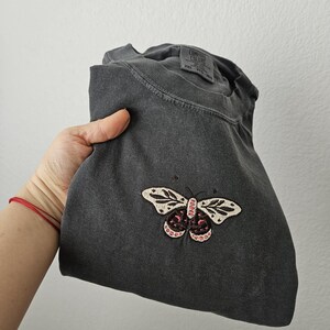 K&ouml;nnte beinhalten: Dunkelgraues T-Shirt mit Schmetterlingsstickerei. Der Schmetterling hat wei&szlig;e Fl&uuml;gel mit schwarzen Details und rosa Akzenten. Das T-Shirt ist gefaltet und wird von einer Hand gehalten.