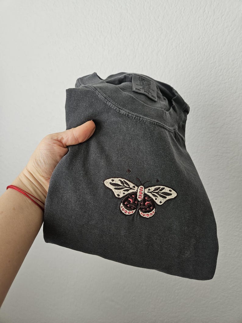 K&ouml;nnte beinhalten: Dunkelgraues T-Shirt mit Schmetterlingsstickerei. Der Schmetterling hat wei&szlig;e Fl&uuml;gel mit schwarzen Details und rosa Akzenten. Das T-Shirt ist gefaltet und wird von einer Hand gehalten.