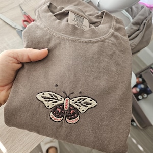 K&ouml;nnte beinhalten: Ein braunes T-Shirt mit einem gestickten Schmetterlingsdesign. Der Schmetterling hat cremefarbene Fl&uuml;gel mit schwarzen Details und einen rosa-schwarzen K&ouml;rper. Das Shirt ist gefaltet und wird von einer Hand gehalten.