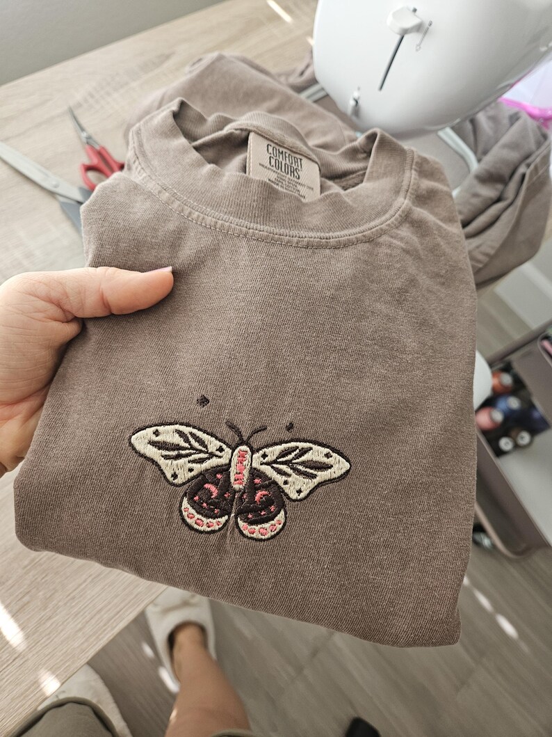 K&ouml;nnte beinhalten: Ein braunes T-Shirt mit einem gestickten Schmetterlingsdesign. Der Schmetterling hat cremefarbene Fl&uuml;gel mit schwarzen Details und einen rosa-schwarzen K&ouml;rper. Das Shirt ist gefaltet und wird von einer Hand gehalten.