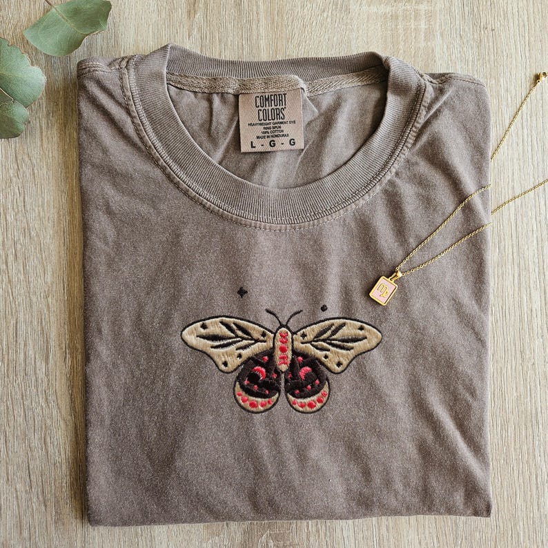 K&ouml;nnte beinhalten: Ein gefaltetes, braunes Comfort Colors T-Shirt mit einem gestickten Motten-Design in Beige, Schwarz und Rot. Eine goldene Halskette mit einem rosa quadratischen Anh&auml;nger liegt auf dem Shirt. Das Shirt-Etikett lautet "Comfort Colors".