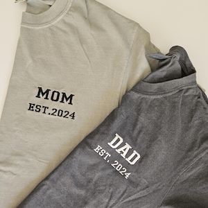 May include: Two t-shirts, one light gray and one dark gray, with the words "MOM EST. 2024" and "DAD EST. 2024" embroidered on them.