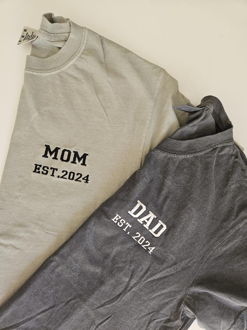 May include: Two t-shirts, one light gray and one dark gray, with the words "MOM EST. 2024" and "DAD EST. 2024" embroidered on them.