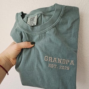May include: A sage green t-shirt with the word "GRANDPA" embroidered in a light tan thread, along with "EST. 2026". The shirt has a crew neck and a tag that reads "COMFORT COLORS".