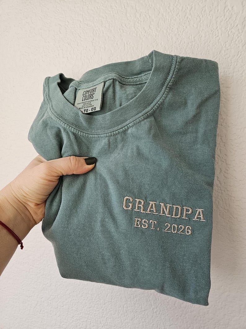 May include: A sage green t-shirt with the word "GRANDPA" embroidered in a light tan thread, along with "EST. 2026". The shirt has a crew neck and a tag that reads "COMFORT COLORS".