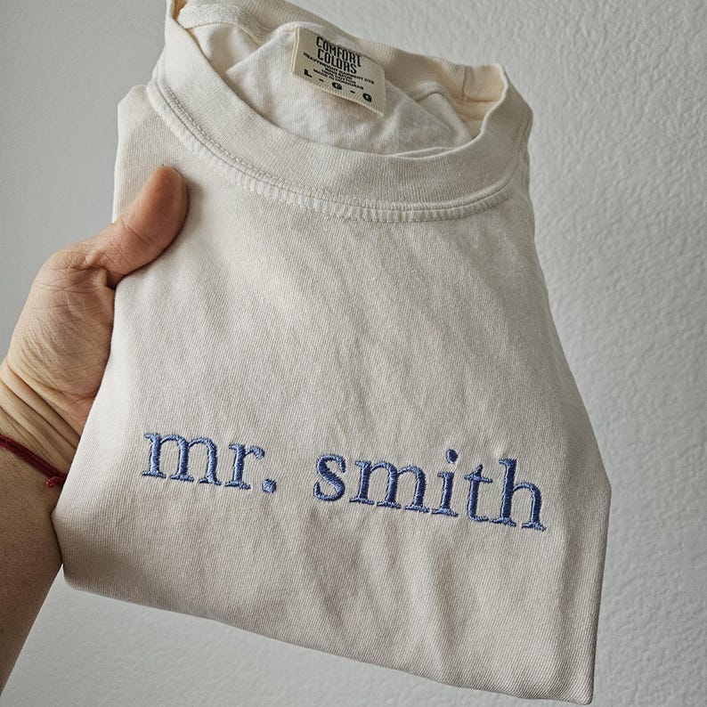 May include: Cream-colored t-shirt with the embroidered text "mr. smith" in blue. The shirt is folded and held by a hand. The tag reads "Comfort Colors".