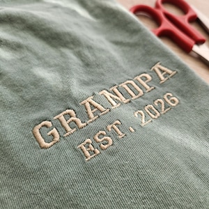 May include: Close-up of a sage green fabric with the embroidered text "GRANDPA EST. 2026" in a light tan color. Red scissors are slightly visible in the background.