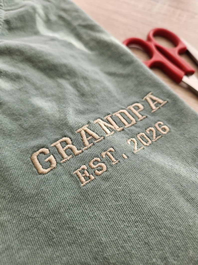 May include: Close-up of a sage green fabric with the embroidered text "GRANDPA EST. 2026" in a light tan color. Red scissors are slightly visible in the background.
