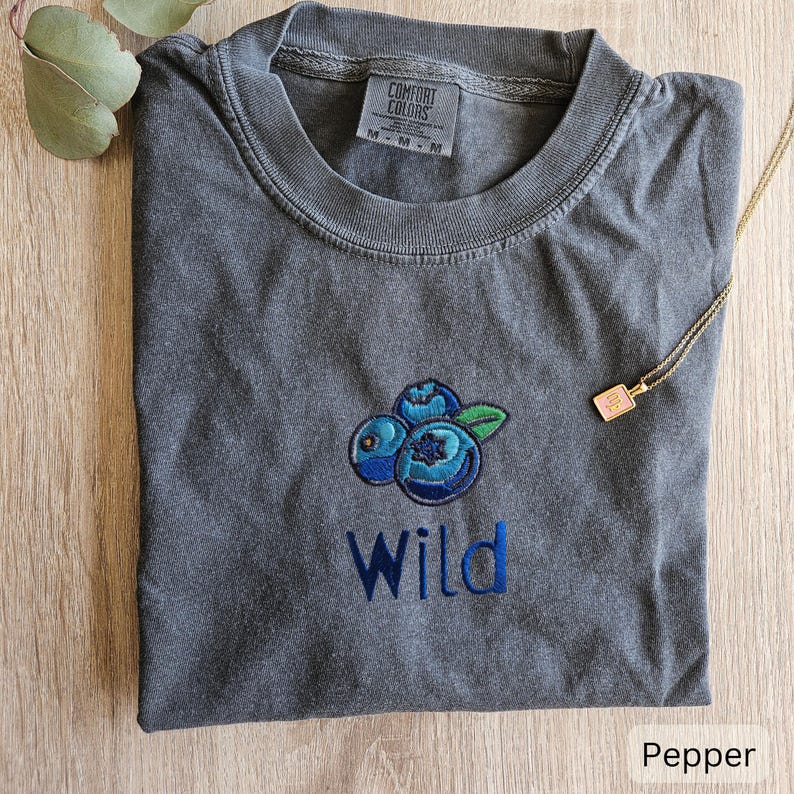 May include: Grey t-shirt with a blue embroidered blueberry design and the word "Wild" underneath. The shirt is made of a soft, comfortable fabric.