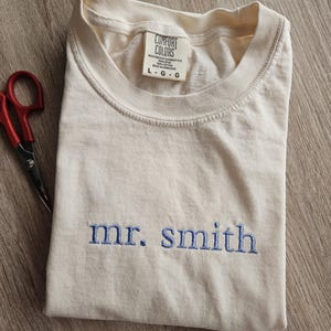 May include: Cream-colored t-shirt with the embroidered text "mr. smith" in blue. The shirt is folded and has a tag that reads "Comfort Colors". A pair of red scissors is visible in the corner.