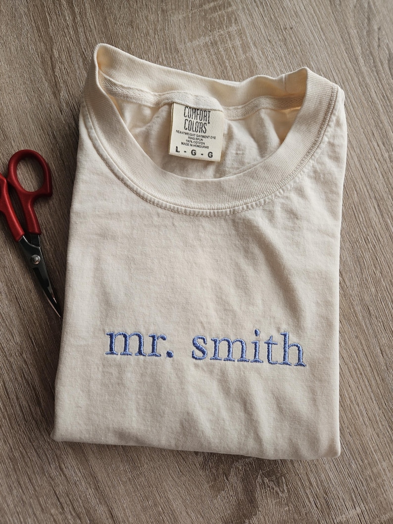 May include: Cream-colored t-shirt with the embroidered text "mr. smith" in blue. The shirt is folded and has a tag that reads "Comfort Colors". A pair of red scissors is visible in the corner.
