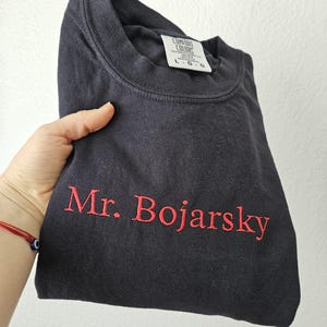 May include: A navy blue t-shirt with the embroidered text "Mr. Bojarsky" in red. The shirt is folded and held in a hand. The tag reads "Comfort Colors" and "L-G-G".