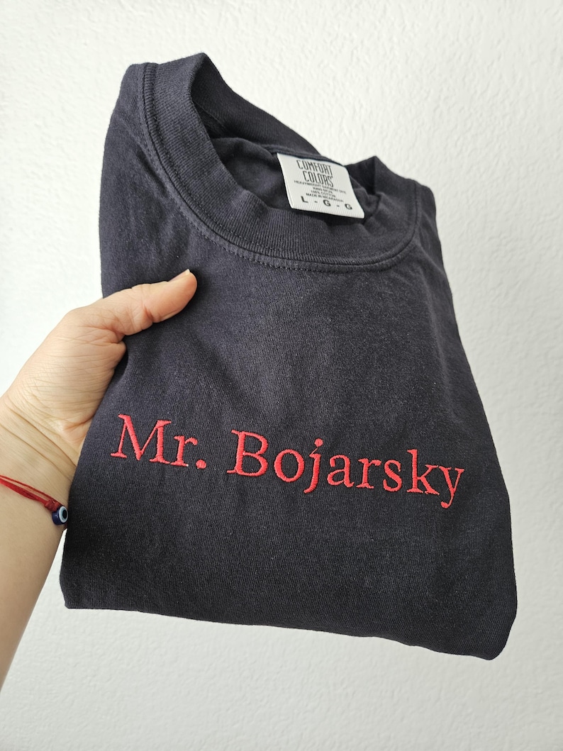 May include: A navy blue t-shirt with the embroidered text "Mr. Bojarsky" in red. The shirt is folded and held in a hand. The tag reads "Comfort Colors" and "L-G-G".