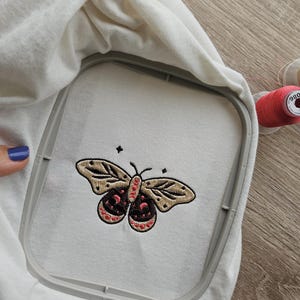 K&ouml;nnte beinhalten: Ein wei&szlig;er Stoff mit einem gestickten Schmetterlingsdesign in Beige, Schwarz und Rot, gehalten in einem Stickrahmen. Der Schmetterling hat detaillierte Fl&uuml;gelmuster und ein symmetrisches Design. Eine Spule rosa Garn ist im Hintergrund sichtbar.
