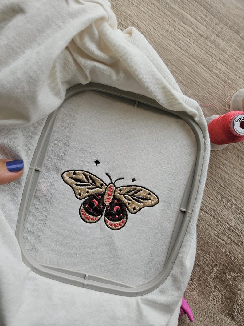 K&ouml;nnte beinhalten: Ein wei&szlig;er Stoff mit einem gestickten Schmetterlingsdesign in Beige, Schwarz und Rot, gehalten in einem Stickrahmen. Der Schmetterling hat detaillierte Fl&uuml;gelmuster und ein symmetrisches Design. Eine Spule rosa Garn ist im Hintergrund sichtbar.