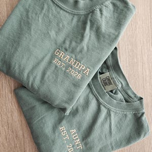 May include: Two sage green Comfort Colors t-shirts. One shirt has "GRANDPA EST. 2026" embroidered in cream thread. The other shirt has "AUNT EST. 2026" embroidered in cream thread. Both shirts are folded and displayed on a light wood surface.