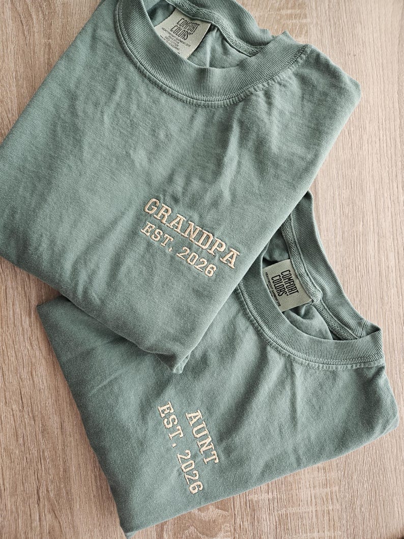 May include: Two sage green Comfort Colors t-shirts. One shirt has "GRANDPA EST. 2026" embroidered in cream thread. The other shirt has "AUNT EST. 2026" embroidered in cream thread. Both shirts are folded and displayed on a light wood surface.