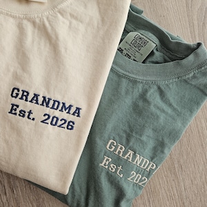 May include: Two Comfort Colors t-shirts, one cream and one teal. The cream shirt has "GRANDMA Est. 2026" embroidered in navy blue. The teal shirt has "GRANDPA Est. 202" embroidered in white. Both shirts have the Comfort Colors tag.