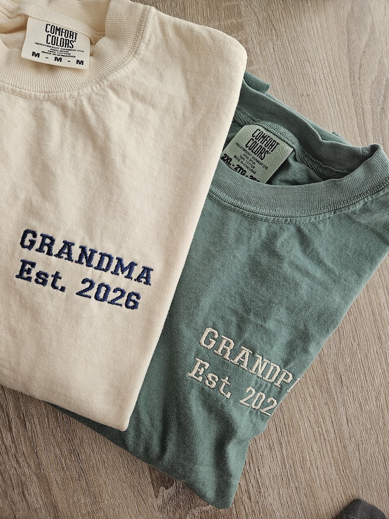 May include: Two Comfort Colors t-shirts, one cream and one teal. The cream shirt has "GRANDMA Est. 2026" embroidered in navy blue. The teal shirt has "GRANDPA Est. 202" embroidered in white. Both shirts have the Comfort Colors tag.