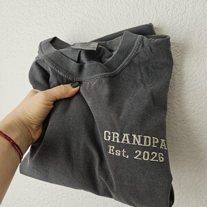 May include: Dark gray t-shirt with the embroidered text "GRANDPA Est. 2026". The shirt is folded and held by a hand. The shirt has a crew neck and appears to be made of a soft, comfortable material.