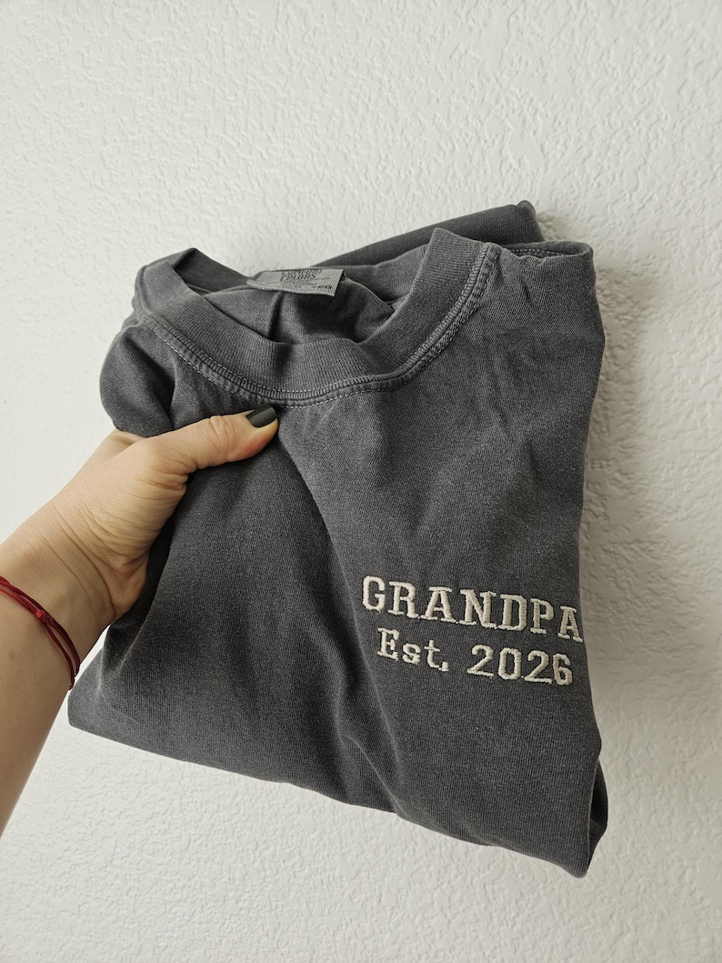 May include: Dark gray t-shirt with the embroidered text "GRANDPA Est. 2026". The shirt is folded and held by a hand. The shirt has a crew neck and appears to be made of a soft, comfortable material.