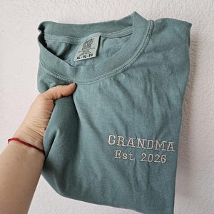 May include: A folded, seafoam green t-shirt with the embroidered text "GRANDMA Est. 2026" in white thread. The shirt has a crew neck and a tag that reads "COMFORT COLORS". The shirt is a size XL.