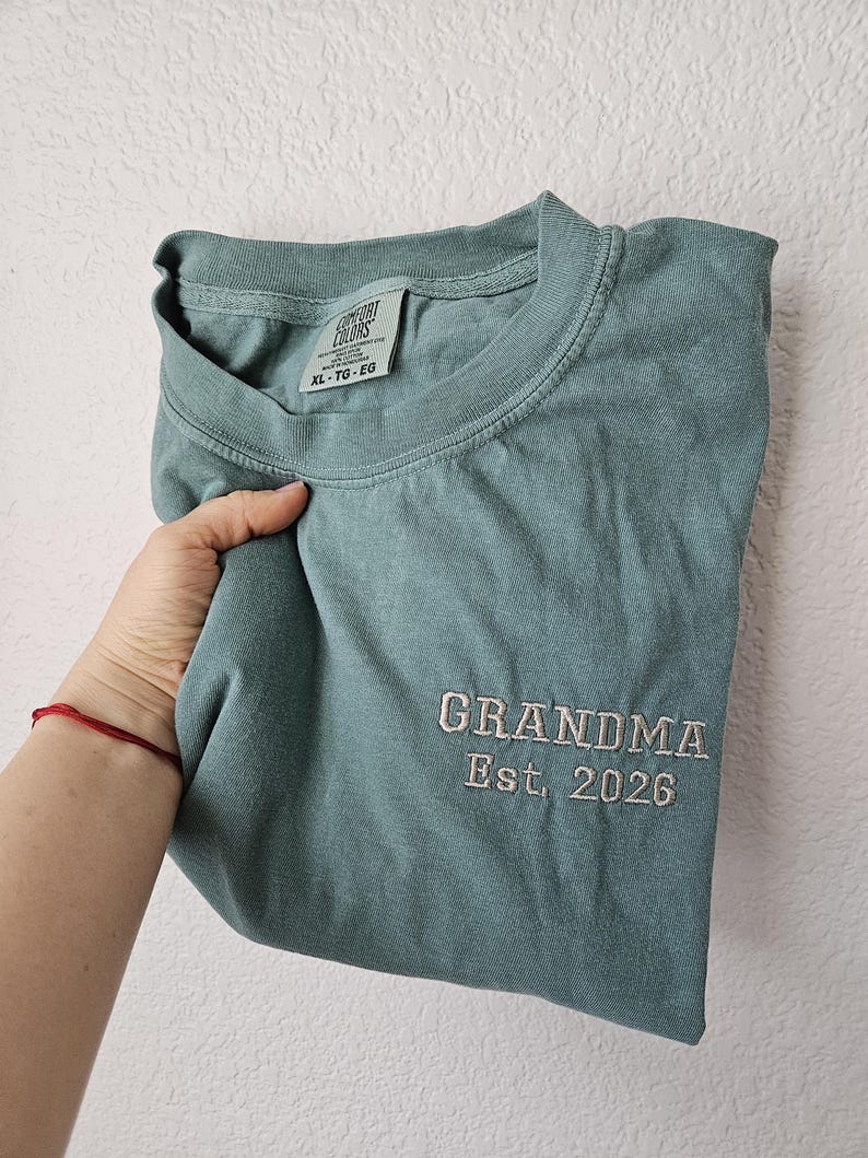 May include: A folded, seafoam green t-shirt with the embroidered text "GRANDMA Est. 2026" in white thread. The shirt has a crew neck and a tag that reads "COMFORT COLORS". The shirt is a size XL.