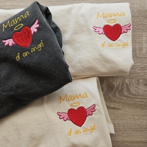 Puede incluir: Tres camisetas blancas con el texto "Mama of an angel" bordado en amarillo con un coraz&oacute;n rojo y alas rosas sobre el texto. El coraz&oacute;n tiene un halo amarillo encima.