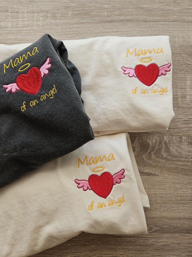 Puede incluir: Tres camisetas blancas con el texto "Mama of an angel" bordado en amarillo con un coraz&oacute;n rojo y alas rosas sobre el texto. El coraz&oacute;n tiene un halo amarillo encima.