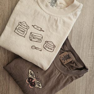 K&ouml;nnte beinhalten: Zwei T-Shirts, eines cremefarben und eines braun, werden gezeigt. Das cremefarbene Shirt zeigt gestickte Backmotive: Mehl, Nudelholz, Butter, Zucker und einen Schneebesen. Das braune Shirt hat ein Schmetterlingsdesign. Beide Shirts haben das "Comfort Colors"-Etikett.