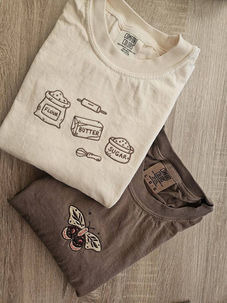 K&ouml;nnte beinhalten: Zwei T-Shirts, eines cremefarben und eines braun, werden gezeigt. Das cremefarbene Shirt zeigt gestickte Backmotive: Mehl, Nudelholz, Butter, Zucker und einen Schneebesen. Das braune Shirt hat ein Schmetterlingsdesign. Beide Shirts haben das "Comfort Colors"-Etikett.