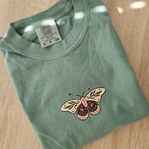 K&ouml;nnte beinhalten: Salbeigr&uuml;nes T-Shirt mit Schmetterlingsstickerei. Der Schmetterling hat beige Fl&uuml;gel mit einem braunen und rosa K&ouml;rper. Das Shirt ist gefaltet und hat ein Etikett mit der Aufschrift "COMFORT COLORS".