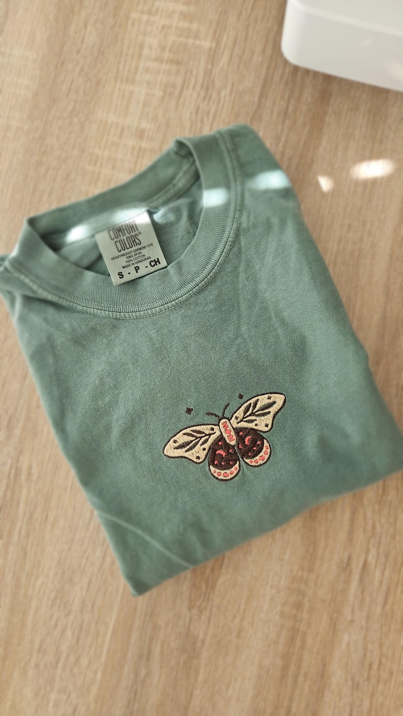 K&ouml;nnte beinhalten: Salbeigr&uuml;nes T-Shirt mit Schmetterlingsstickerei. Der Schmetterling hat beige Fl&uuml;gel mit einem braunen und rosa K&ouml;rper. Das Shirt ist gefaltet und hat ein Etikett mit der Aufschrift "COMFORT COLORS".
