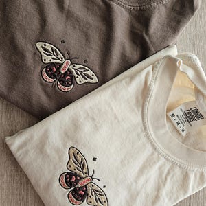 K&ouml;nnte beinhalten: Zwei gefaltete T-Shirts, eines braun und eines cremefarben, jeweils mit einem Schmetterlingsdesign bestickt. Der Schmetterling hat cremefarbene Fl&uuml;gel mit braunen und rosa Akzenten. Die Shirts liegen auf einer Holzoberfl&auml;che.