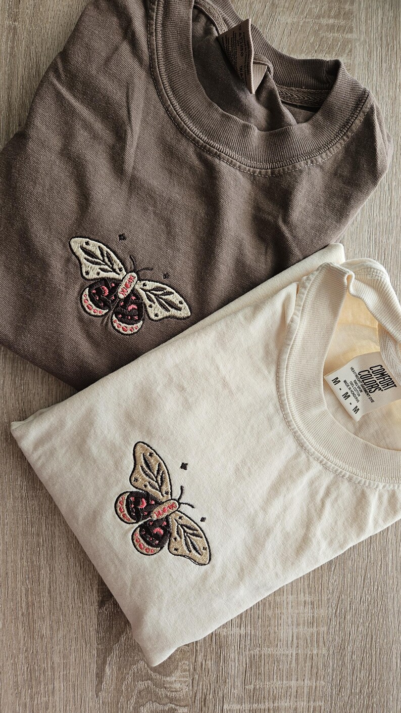 K&ouml;nnte beinhalten: Zwei gefaltete T-Shirts, eines braun und eines cremefarben, jeweils mit einem Schmetterlingsdesign bestickt. Der Schmetterling hat cremefarbene Fl&uuml;gel mit braunen und rosa Akzenten. Die Shirts liegen auf einer Holzoberfl&auml;che.