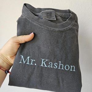 May include: A dark gray t-shirt with the embroidered text "Mr. Kashon" in light blue. The shirt is folded and held by a hand. The tag reads "Comfort Colors".