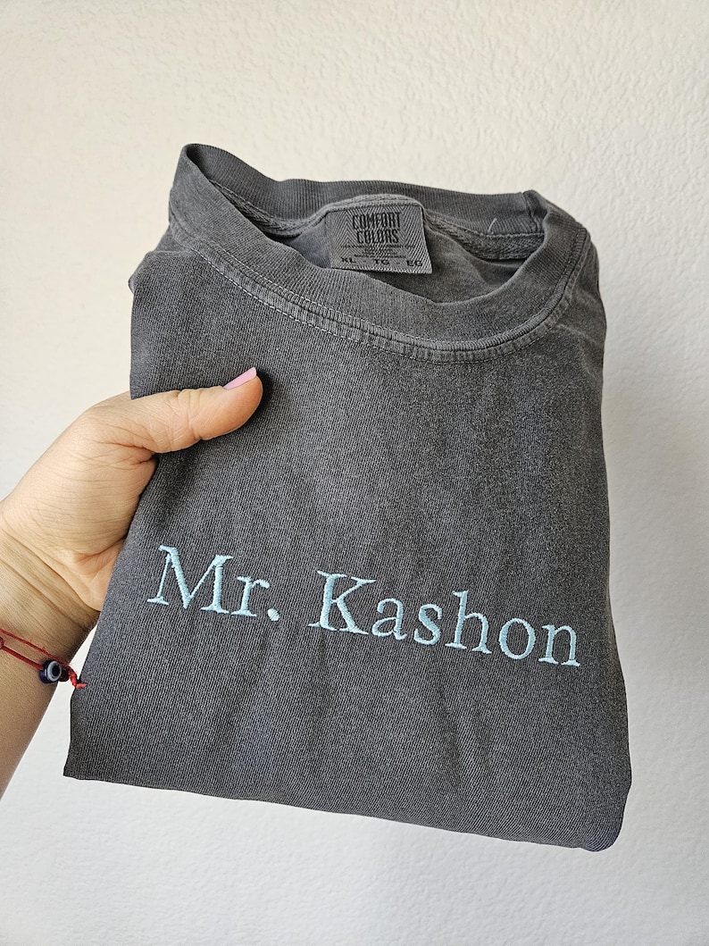 May include: A dark gray t-shirt with the embroidered text "Mr. Kashon" in light blue. The shirt is folded and held by a hand. The tag reads "Comfort Colors".