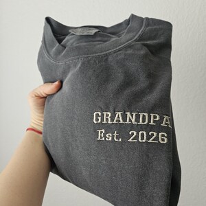 May include: Dark gray sweatshirt with the embroidered text "GRANDPA Est. 2026" in a classic font. The sweatshirt is folded, showing the text on the front. The fabric appears soft and comfortable.
