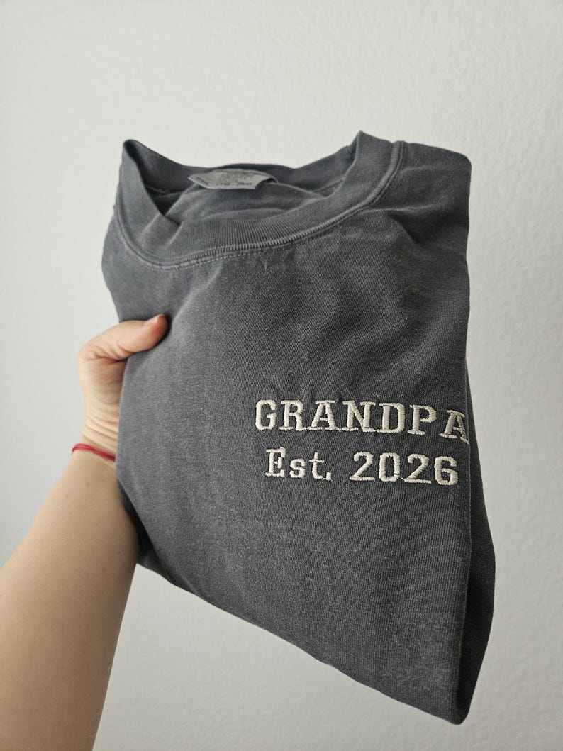 May include: Dark gray sweatshirt with the embroidered text "GRANDPA Est. 2026" in a classic font. The sweatshirt is folded, showing the text on the front. The fabric appears soft and comfortable.