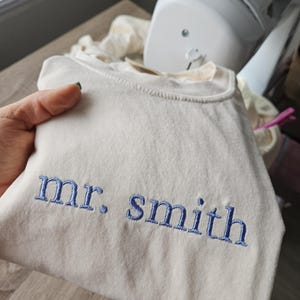 May include: Cream-colored sweatshirt with the embroidered text "mr. smith" in blue thread. The sweatshirt is folded, and a sewing machine is visible in the background.