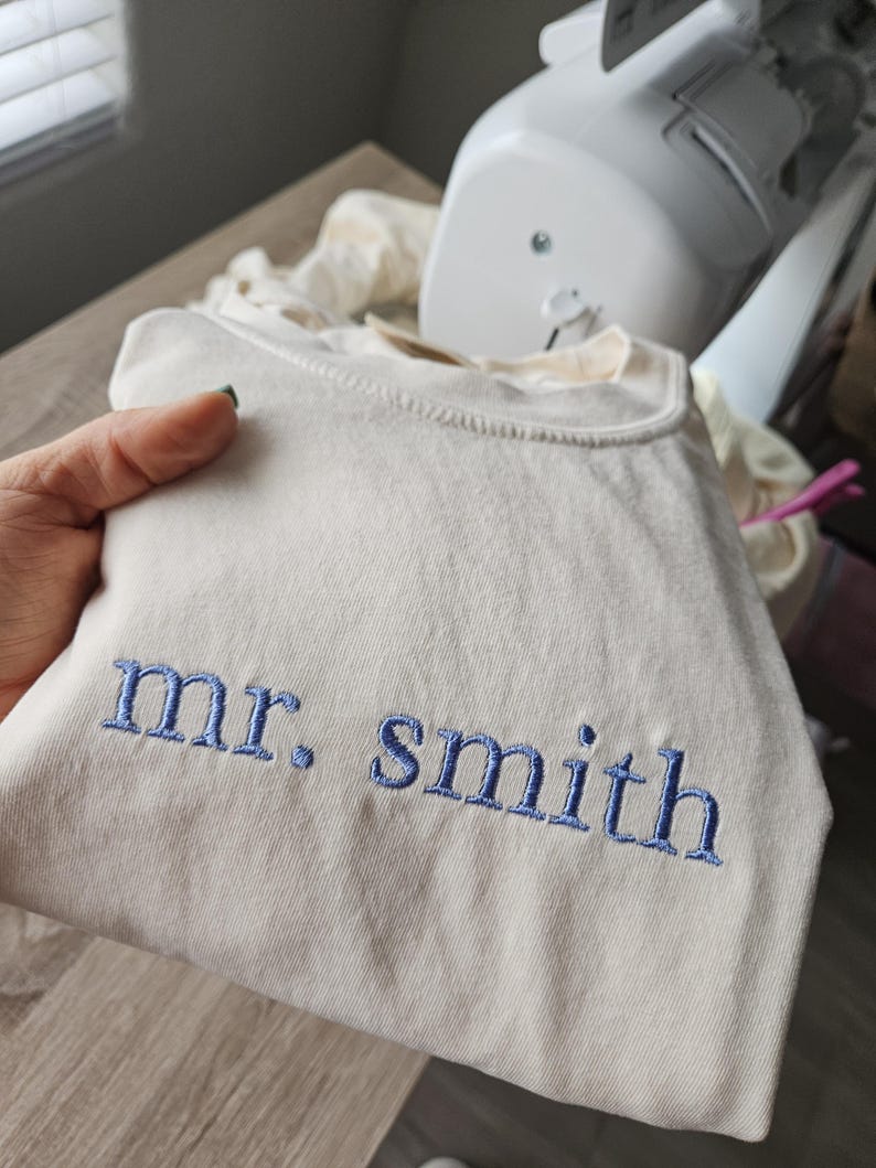 May include: Cream-colored sweatshirt with the embroidered text "mr. smith" in blue thread. The sweatshirt is folded, and a sewing machine is visible in the background.