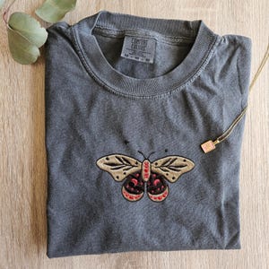 K&ouml;nnte beinhalten: Ein graues Comfort Colors T-Shirt mit einem gestickten Schmetterlingsdesign. Der Schmetterling hat beige Fl&uuml;gel mit schwarzen und roten Akzenten. Das Shirt hat ein kleines Etikett mit der Aufschrift "Comfort Colors". Eine goldene Halskette mit einem kleinen Anh&auml;nger ist ebenfalls sichtbar.