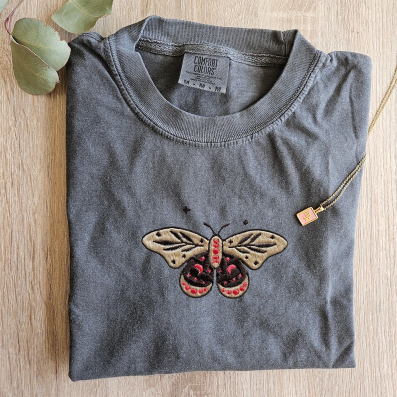 K&ouml;nnte beinhalten: Ein graues Comfort Colors T-Shirt mit einem gestickten Schmetterlingsdesign. Der Schmetterling hat beige Fl&uuml;gel mit schwarzen und roten Akzenten. Das Shirt hat ein kleines Etikett mit der Aufschrift "Comfort Colors". Eine goldene Halskette mit einem kleinen Anh&auml;nger ist ebenfalls sichtbar.