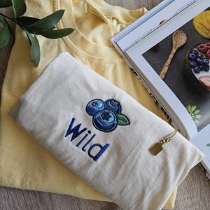 May include: A white t-shirt with a blue embroidered blueberry design and the word "Wild" in blue embroidery.