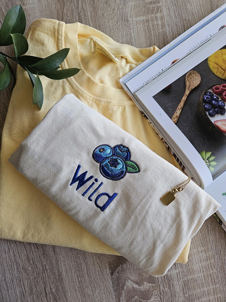 May include: A white t-shirt with a blue embroidered blueberry design and the word "Wild" in blue embroidery.