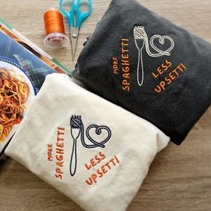 May include: Two cream-colored and dark gray sweatshirts with embroidered spaghetti and heart designs. The text on the sweatshirts reads "More Spaghetti, Less Upsetti".