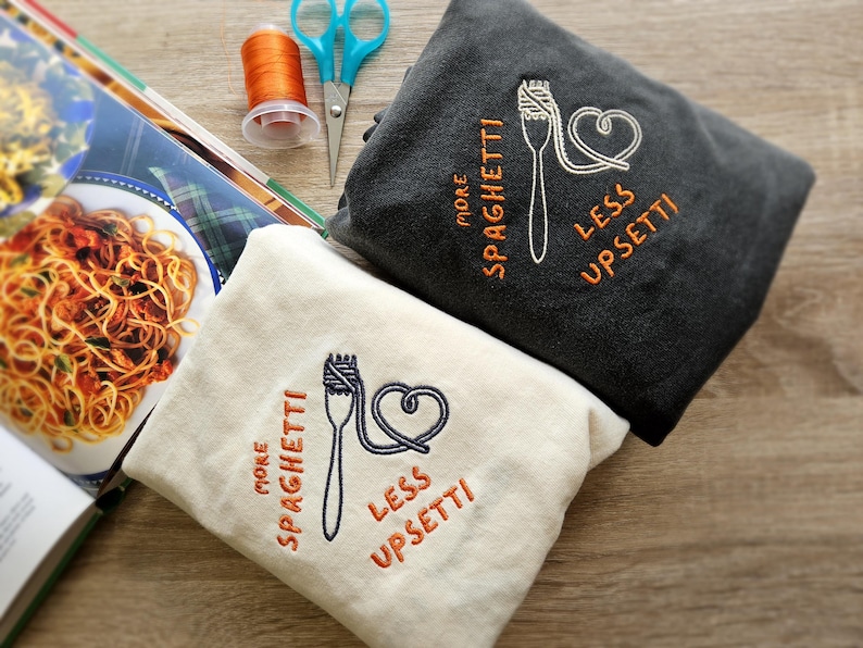 May include: Two cream-colored and dark gray sweatshirts with embroidered spaghetti and heart designs. The text on the sweatshirts reads "More Spaghetti, Less Upsetti".