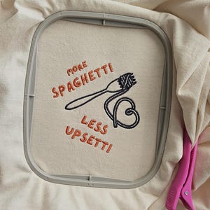 May include: A beige t-shirt with an embroidered design that says "More Spaghetti Less Upsetti!" with a fork and spaghetti noodles.