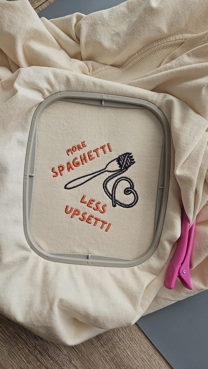 May include: A beige t-shirt with an embroidered design that says "More Spaghetti Less Upsetti!" with a fork and spaghetti noodles.