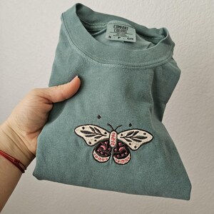 K&ouml;nnte beinhalten: Ein gefaltetes, salbeigr&uuml;nes T-Shirt mit einem gestickten Schmetterlingsdesign. Der Schmetterling hat cremefarbene Fl&uuml;gel mit schwarzen Details und rosa Akzenten. Das Shirt hat den Aufdruck "COMFORT COLORS" auf dem Etikett.